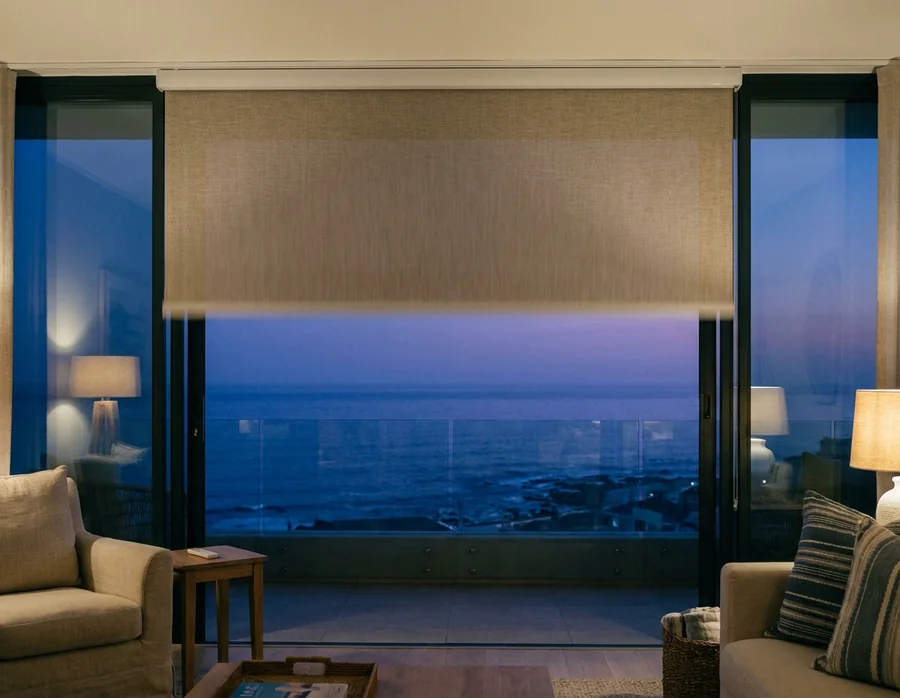 Motorised roller blind paused halfway down wide sliding glass at blue hour, twilight ocean beyond the balcony rail