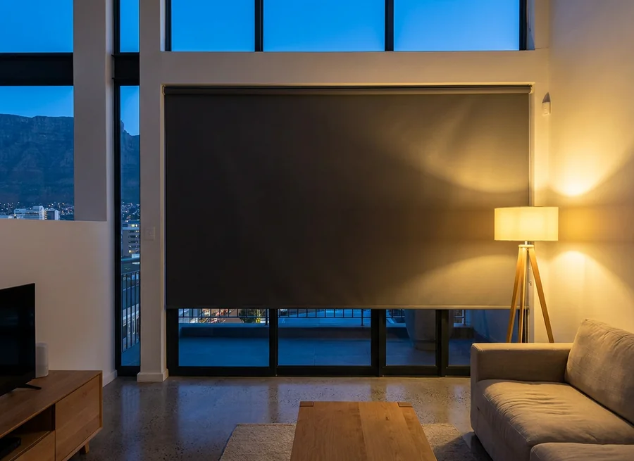 Charcoal blockout roller blind three-quarters down a double-height Cape Town apartment window at dusk, tripod lamp glowing beside it