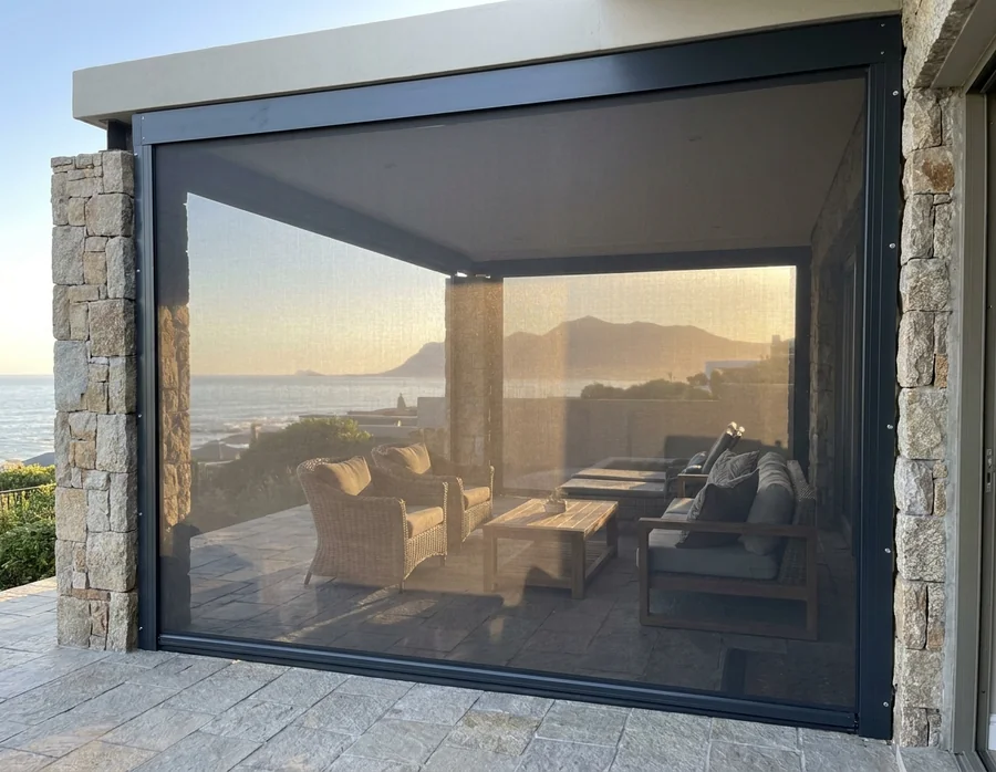 Zip screen mesh held taut in charcoal side channels across a stone-flanked patio opening, evening sea glow behind the weave